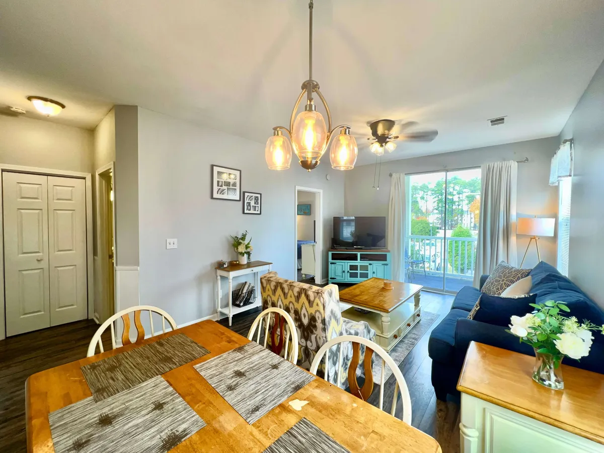 A warmly lit interior featuring a dining table, comfortable sofa, stylish light fixture, and a television. Large windows lead to a balcony, providing a natural connection to the outdoors and letting in ample daylight. The room feels homely and is ideal for gatherings and relaxation.