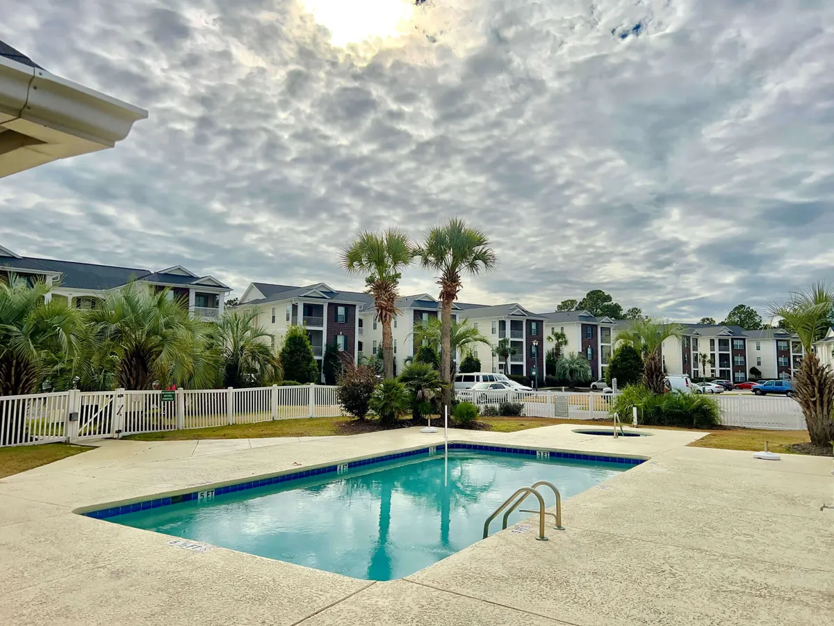 A vibrant outdoor pool area surrounded by palm trees and residential buildings glows in the sunlight. People can be seen swimming, lounging, and socializing, highlighting the lively community spirit and leisure options just steps from the residence.