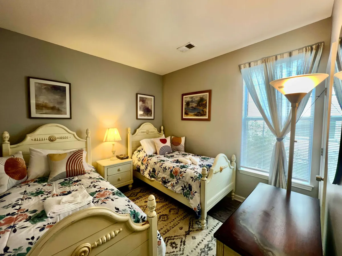 A comfortable bedroom with two beds adorned in floral bedding, a nightstand with a lamp, tasteful artwork on the walls, and a window with sheer curtains letting in soft daylight. The setting radiates comfort and elegant design.