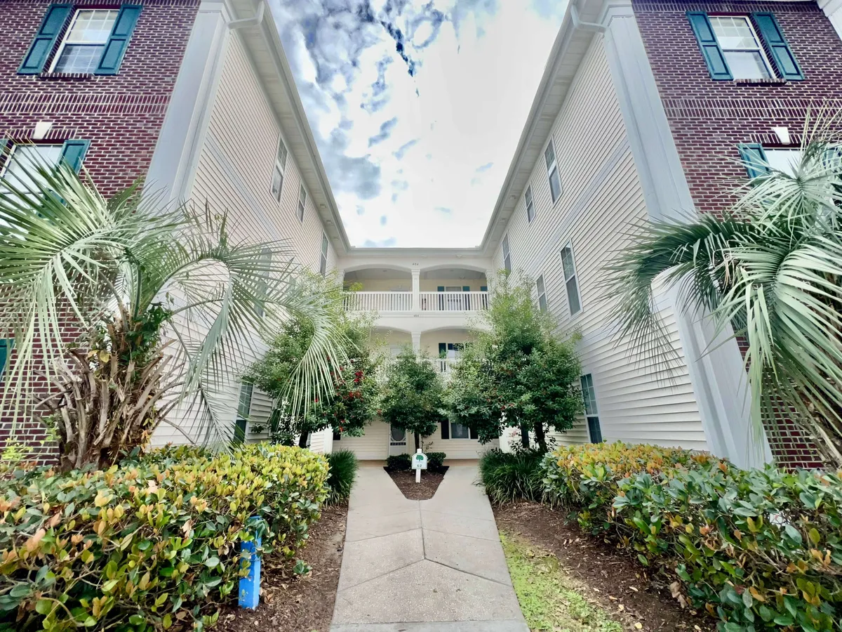 An attractive exterior view of the community, featuring tall palm trees, manicured green shrubs, a sturdy brick facade, and white balconies. The landscaped grounds and inviting outdoor spaces suggest a peaceful yet vibrant community life amidst Myrtle Beach's excitement.