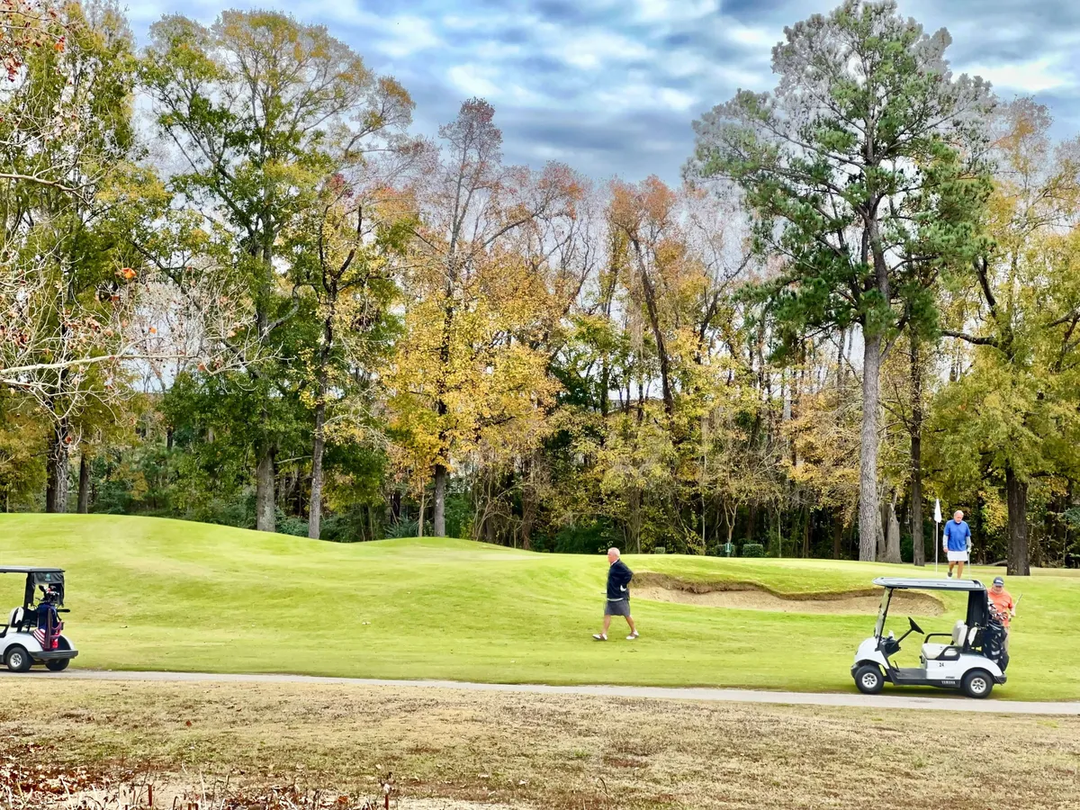 This image captures golfers walking across the course with golf carts nearby, surrounded by trees displaying vibrant fall foliage. The scene is active and lively, highlighting people engaged in the sport and the natural beauty of the course.