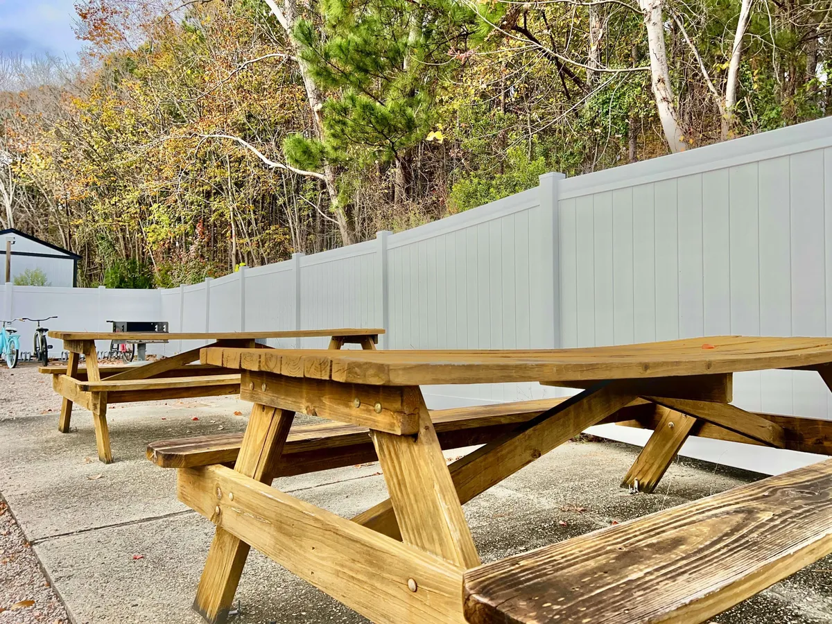 Wooden picnic tables are arranged in a fenced outdoor area beneath leafy trees, perfect for social gatherings, sharing stories, or enjoying the natural environment. The area feels welcoming and communal.