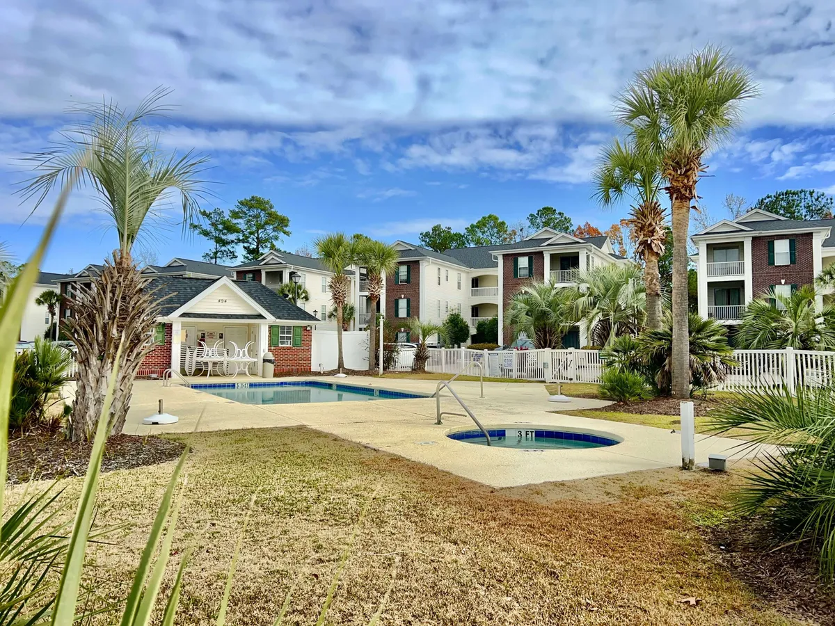 The image showcases the condo's inviting outdoor area, featuring a sparkling swimming pool surrounded by palm trees, plush lounge chairs, and a relaxing spa area. The brick building in the background hints at modern comfort, while the open setup encourages both relaxation and social gatherings under the sun or stars.