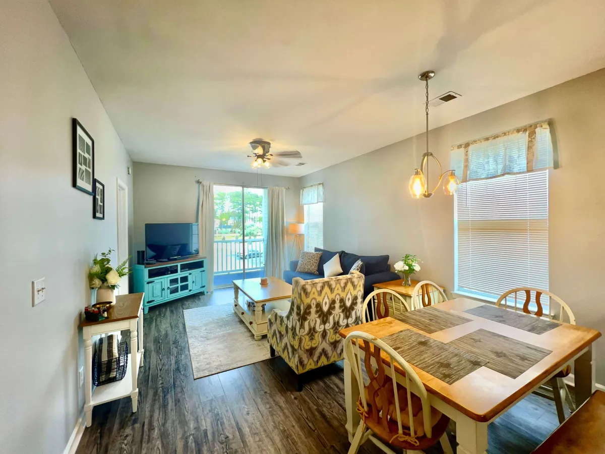 This image showcases a cozy and welcoming living room and dining area. Large windows flood the space with natural light, offering a glimpse of the outdoors. The scene includes a plush sofa, dining table, television, and a decorative plant, highlighting both relaxation and social spaces within the open-plan layout.