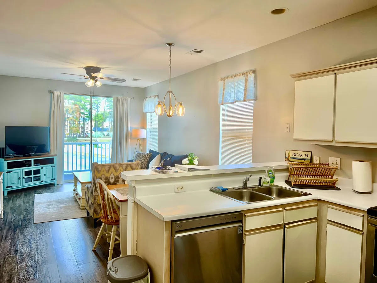 A view of an open-plan kitchen and living area, with a kitchen counter, comfortable couch, television, and dining table all within a single, cohesive space. The layout is modern and inviting, facilitating easy movement and conversation.
