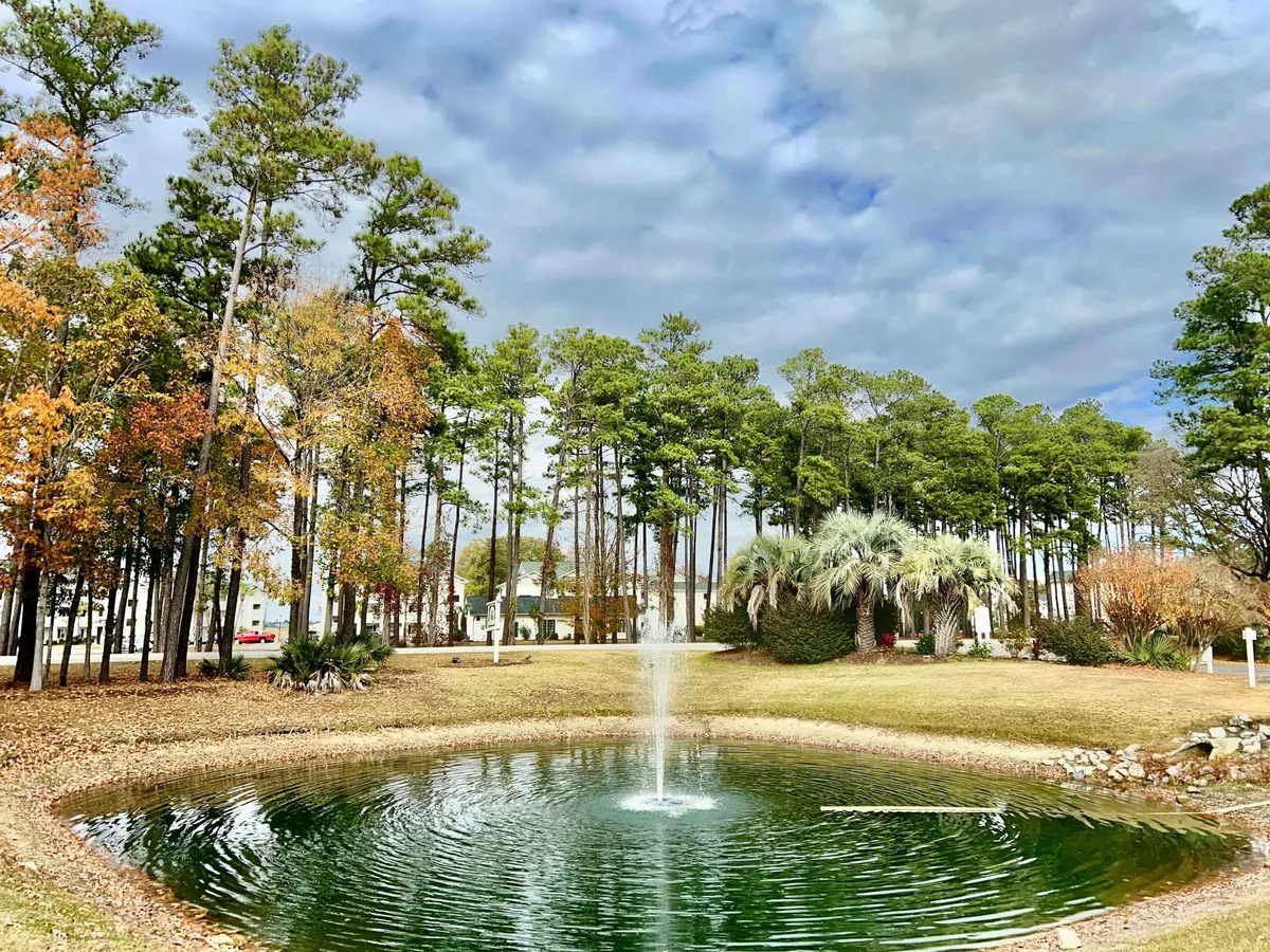 This image shows a tranquil pond with a graceful fountain, surrounded by well-manicured trees and landscaping. The scene is bathed in warm sunset light, with reflections shimmering on the water and a backdrop of autumn foliage. The setting encourages relaxation and invites quiet contemplation of nature.