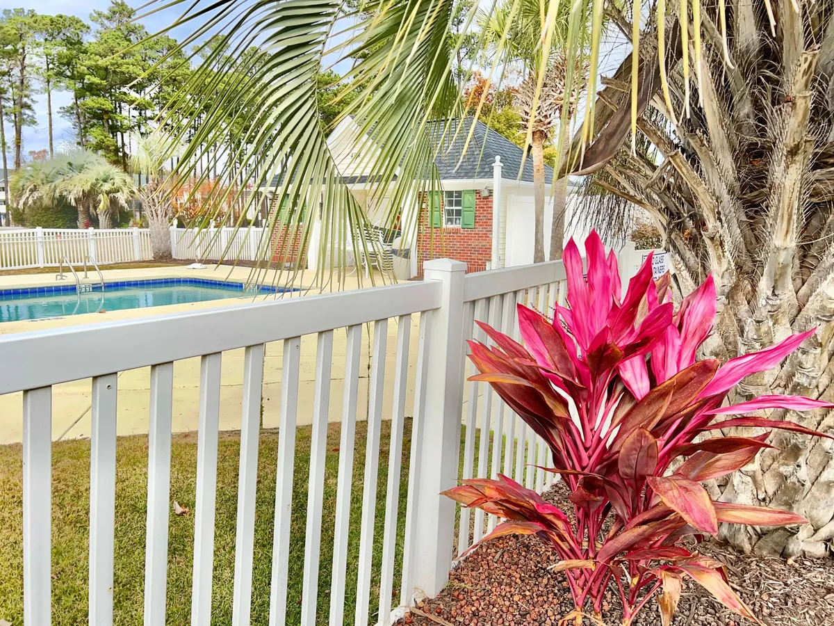 A vibrant exterior pool area surrounded by lush palm trees, bright pink flowering plants, and a crisp white fence. The inviting swimming pool is set against a backdrop of sunset light, with plenty of space for relaxation and socializing, evoking the leisurely charm of a Myrtle Beach getaway.