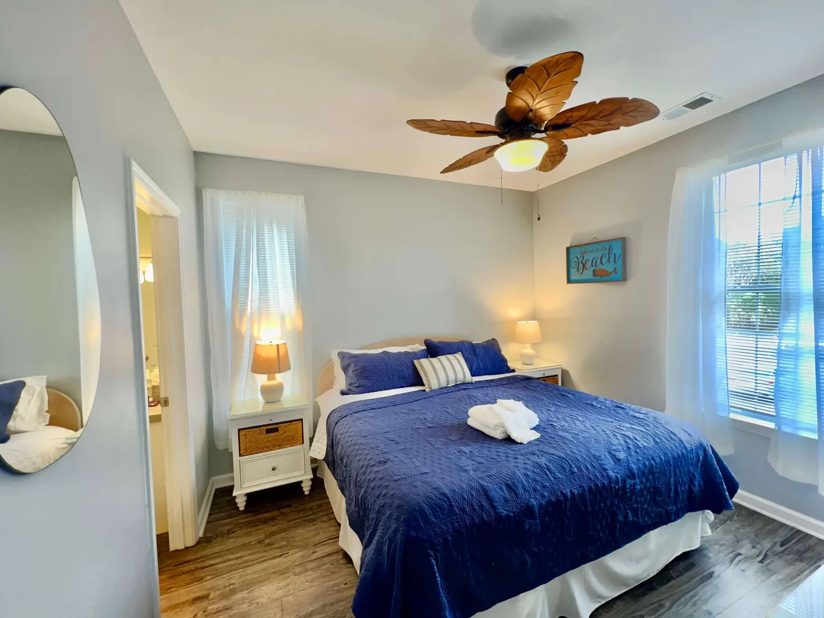 A tranquil bedroom featuring a bed dressed in a blue quilt, a ceiling fan, a nightstand with a lamp, and a framed beach-themed sign on the wall. Gentle natural light fills the room, creating a restful and inviting atmosphere.
